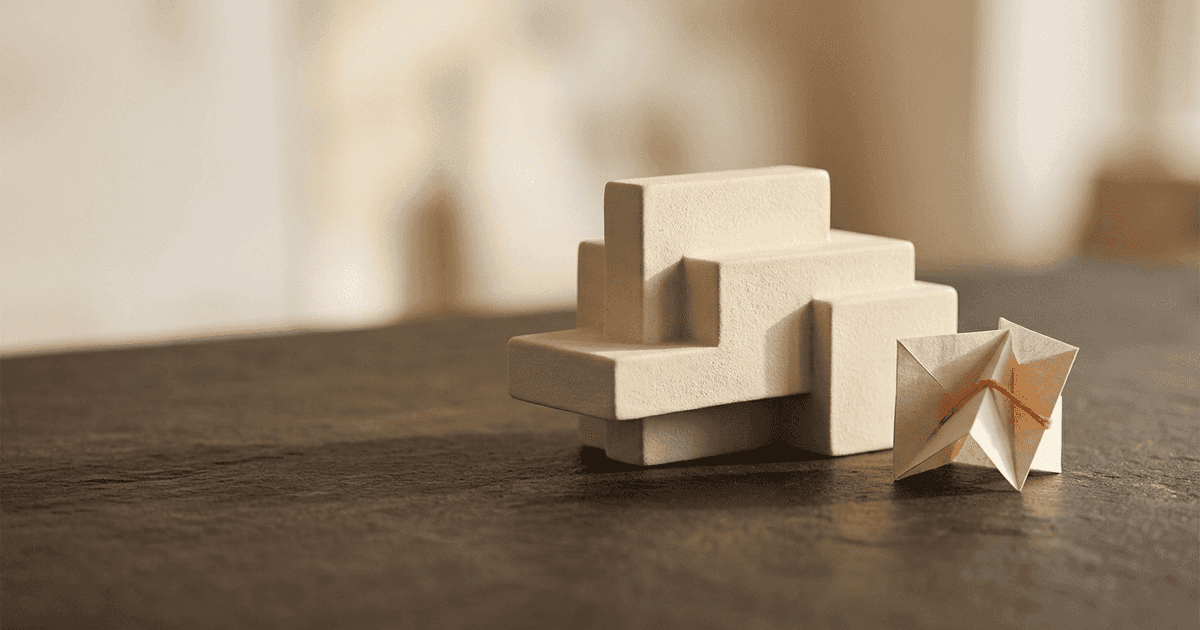 Abstract editorial still life for normalised EBITDA: a cream stepped block form on a dark slate surface beside a small folded paper shape tied with orange thread, suggesting earnings rebuilt step by step